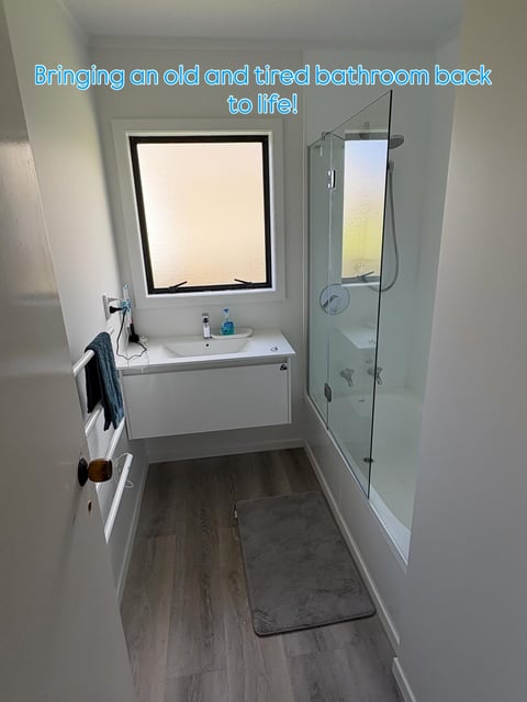 Renovated modern bathroom with white walls, floating vanity, large framed mirror, glass shower enclosure, and wood-look flooring