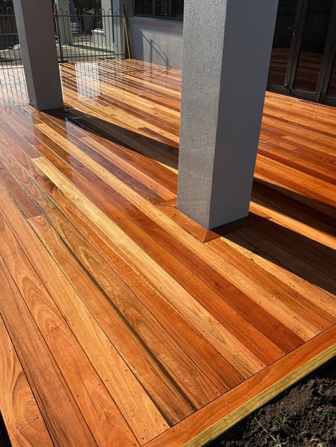 Wooden deck with warm-toned planks and gray support columns, featuring shadows from railings and a gate in the background