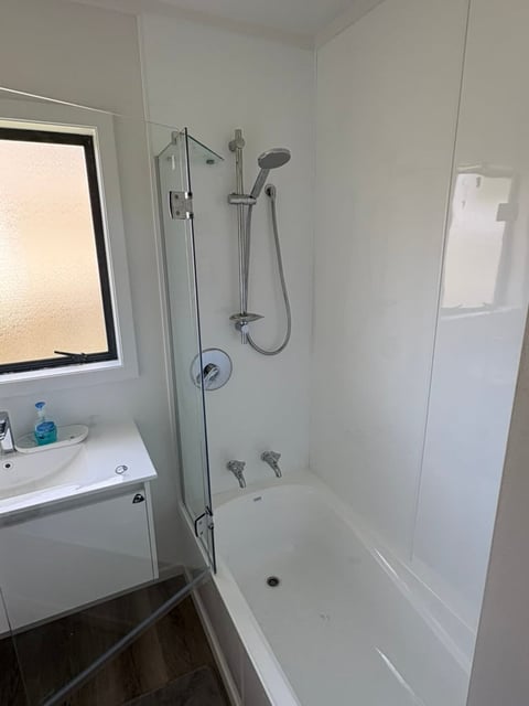 Modern bathroom with white bathtub, glass shower door, chrome fixtures, small sink with mirror, and frosted window