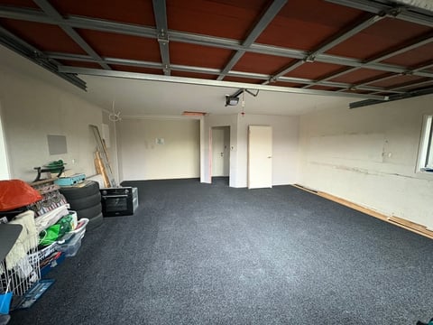 Empty basement room with dark gray carpet flooring, white walls, exposed red wooden ceiling beams, and construction materials stacked in the corner