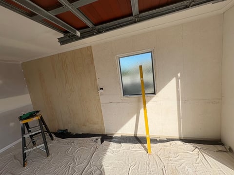 Interior of unfinished room under construction with plywood wall, drop cloth flooring, step ladder, and yellow support post