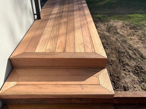 Wooden deck stairs with two steps leading down to bare ground next to white building wall
