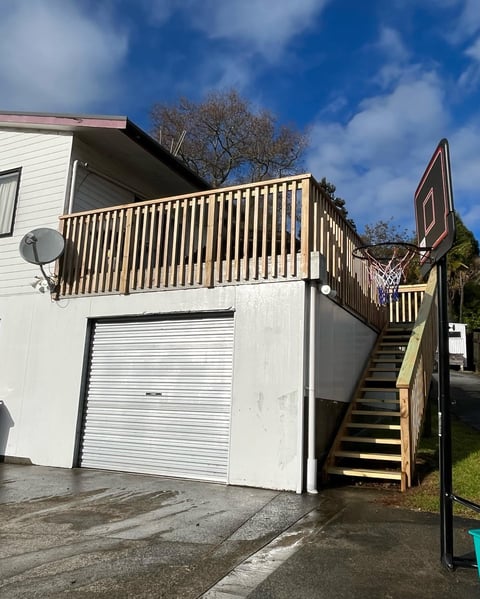 White house with attached garage below an elevated deck with wooden railings and exterior stairs, set against a blue sky