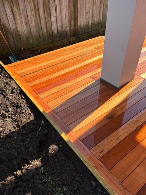 Wooden deck with warm orange-toned planks, gray column support, and dark fence in background with afternoon sunlight casting shadows