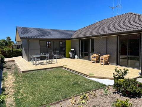 Modern single-story house with wooden deck, outdoor dining area, and manicured lawn under blue sky