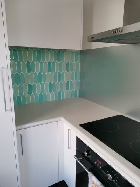Modern kitchen with turquoise geometric patterned backsplash, white countertop, black electric stovetop, and white cabinetry