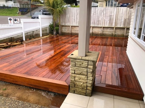 Wooden deck with stained finish, support post, and glass railing overlooking fenced backyard with white house visible