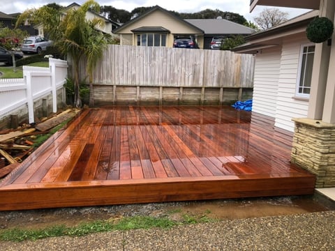 Large wooden deck with reddish-brown stain in backyard with white fence, house, and surrounding trees