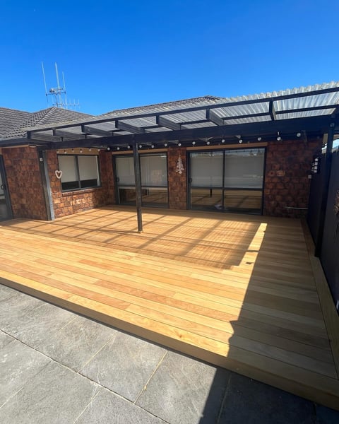Modern timber deck with pergola structure attached to brick house with large windows and clear blue sky