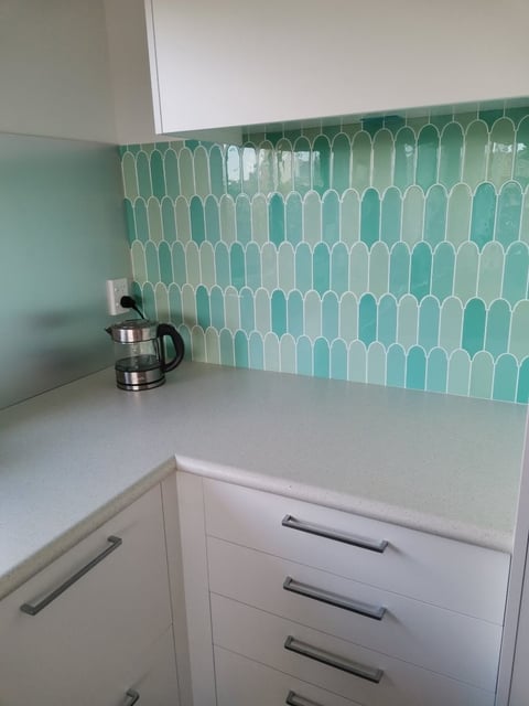 Modern kitchen corner with white cabinetry, turquoise geometric patterned tile backsplash, and electric kettle on white countertop